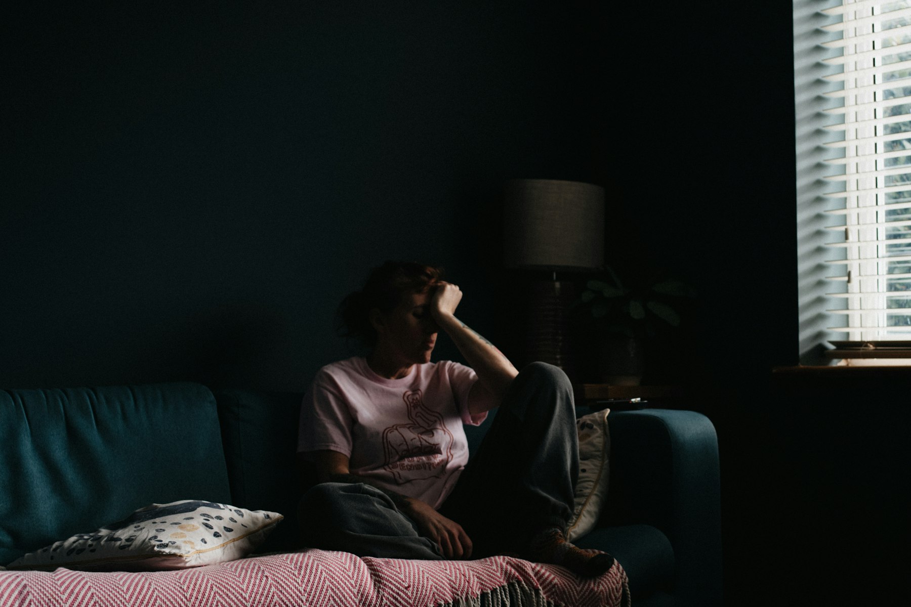 Eine Frau sitzt in einem abgedunkelten Wohnzimmer auf dem Sofa und stützt den Kopf in die Hand